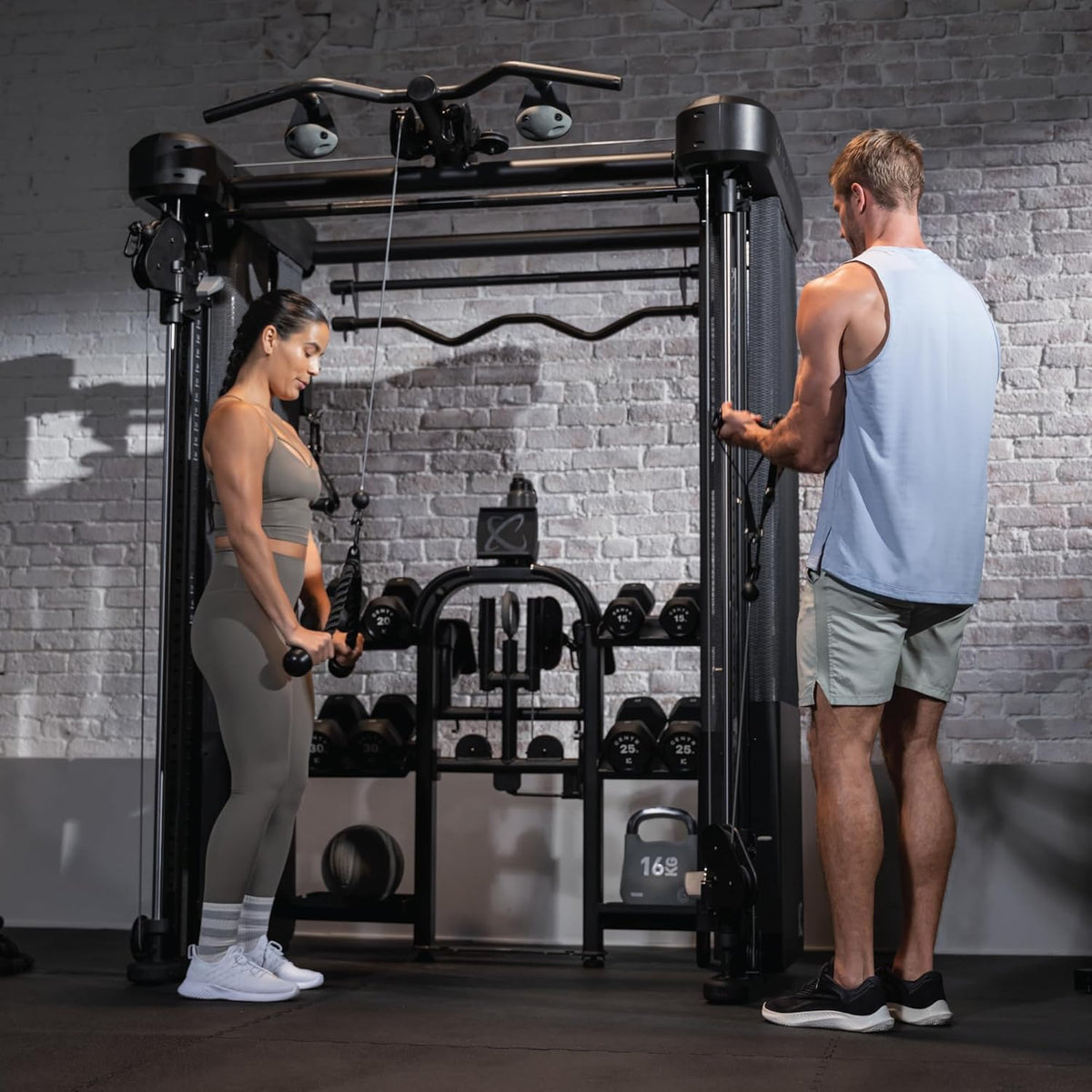Two people using a cable machine in a gym setting with a brick wall background.